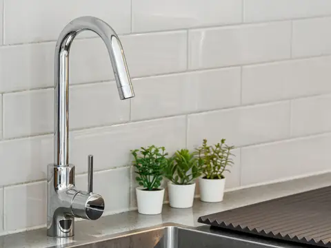 A chrome kitchen faucet against a white tiled wall with small potted plants