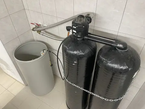 A twin-tank water softener with its brine tank beside it against a tiled wall